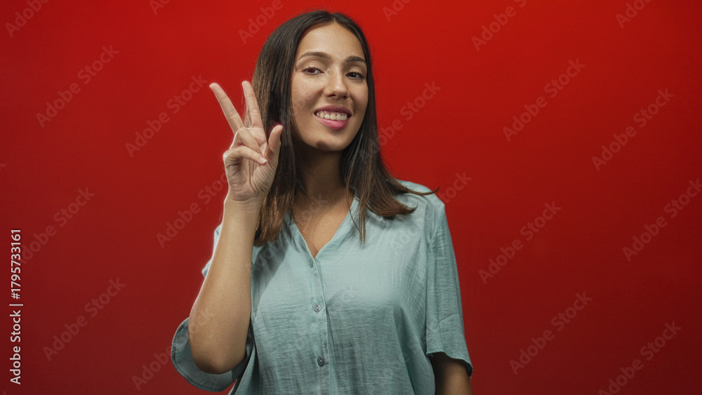 Fototapeta premium Hispanic young brunette woman smiling and showing a peace sign with her hand and visible forearm in a red studio setting; playful.
