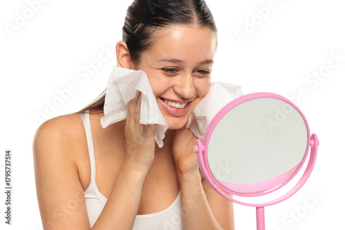 Young smiling woman with long dark hair cleaning her face with wipes in front of a mirror, studio shot on a white background