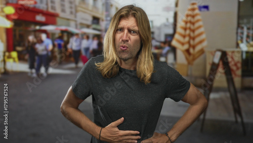 Photos Blond man with long hair visibly grimaces as he clutches stomach on street; pain discomfort nausea distress