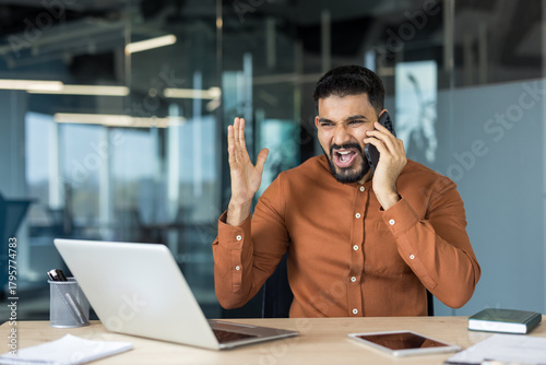 Businessman in modern office yelling into mobile phone while angrily gesturing at laptop, stressed and frustrated during a heated work call and urgent conflict at his desk