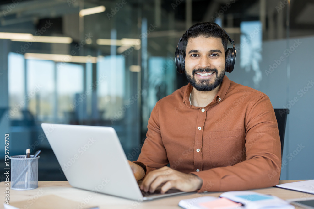 Obraz premium Young indian man in a modern office wearing headphones, smiling at camera while typing on a laptop, engaging in remote work, online learning, or virtual communication