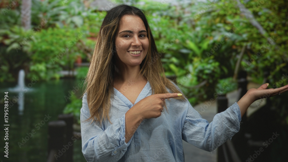 Obraz premium Woman smiling with palm up hand and pointing finger on sunlit forest walkway near pond in green park; joyful welcome.