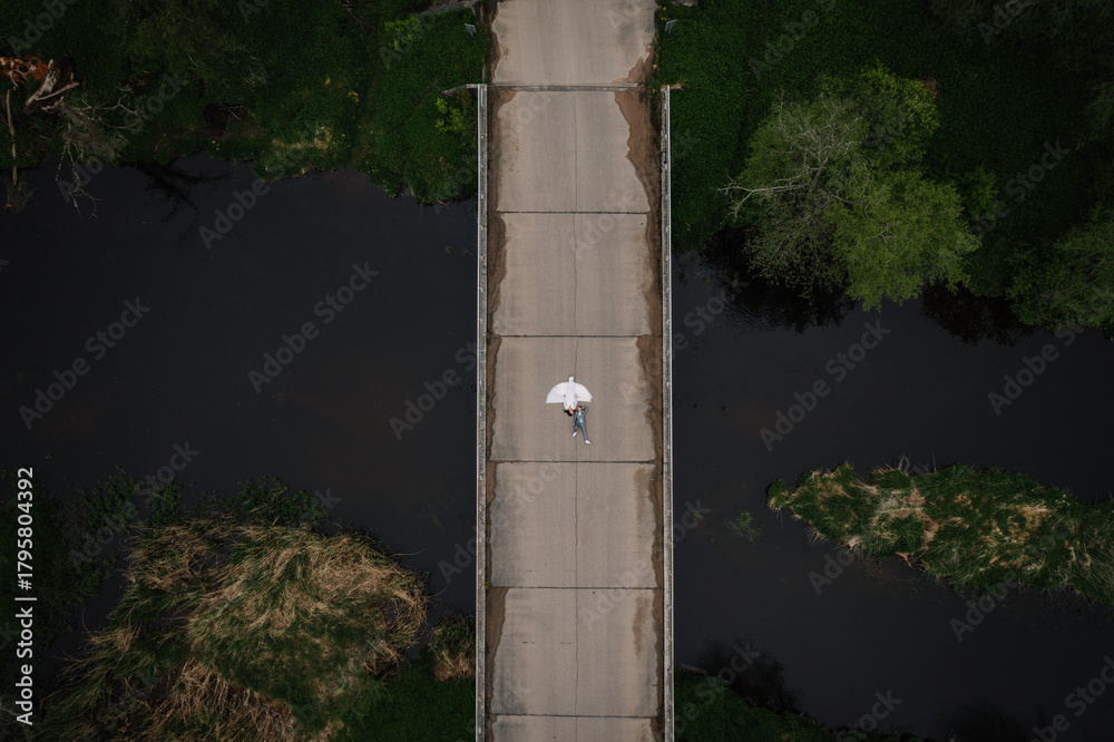 Fototapeta premium Aerial view of a solitary figure walking on a concrete bridge surrounded by lush greenery and dark water, capturing a serene moment in nature's embrace