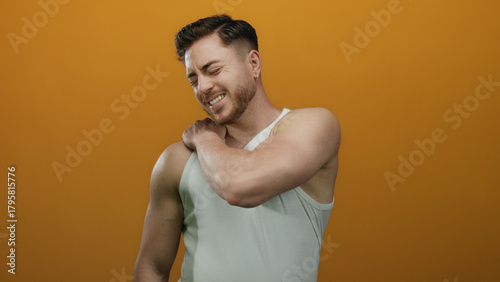 Schilderij op canvas Young man with beard in white tank top experiencing shoulder pain against isolated orange background, conveying discomfort and health theme