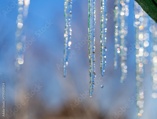 row of shiny cold icicles hang on a green roof on a sunny spring day against ...