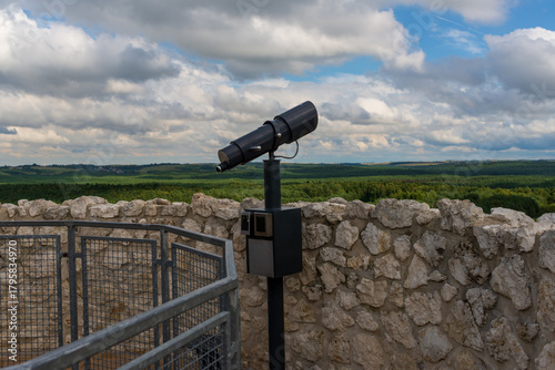 A metal viewing telescope stands on a stone platform above a vast rural panorama. Rolling forests, open fields and dramatic clouds create an inspiring scenic viewpoint.