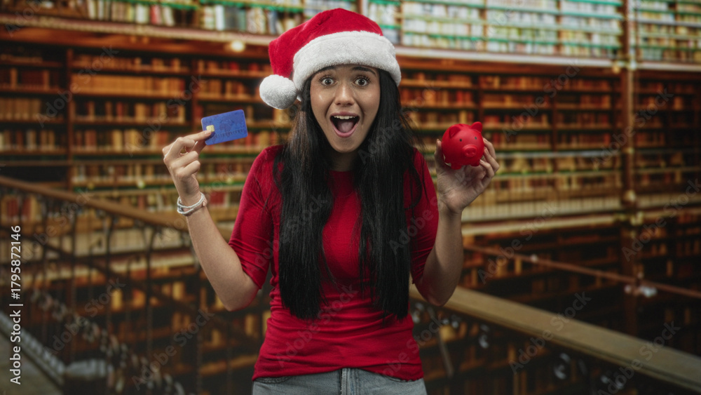 Fototapeta premium Woman holding a creditcard in one hand and a red piggybank in the other with a surprised expression in a building; holiday budgeting concern.