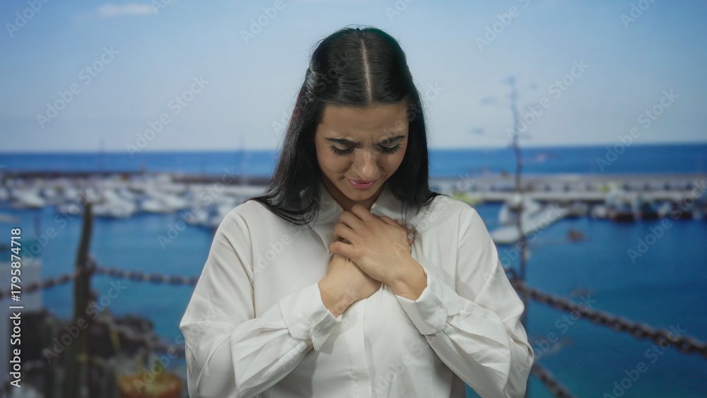 Fototapeta premium Woman with hands on chest standing at a port with boats in the background, wearing a white shirt, demonstrating an emotional and reflective moment outdoors by the sea.