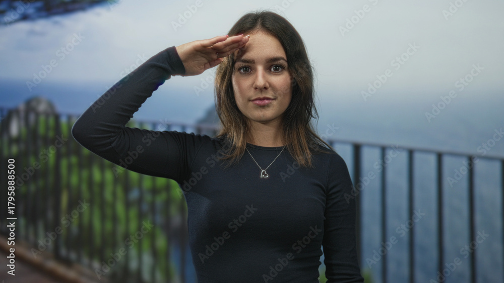 Obraz premium Young woman salutes with hand to forehead on building terrace by railing overlooking ocean view; confidence.