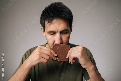 Studio portrait of enjoying young man savoring rich aroma of dark chocolate bar with closed eyes, holding it close to face in anticipation of delightful treat, sitting at table on isolated background.