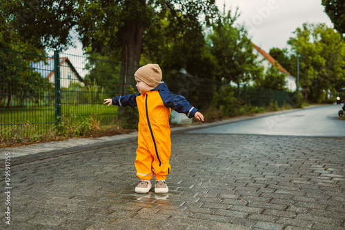 Little child in bright orange rain gear plays joyfully on a cobbled street surrounded by greenery on a cloudy day