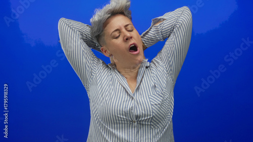 Woman in striped shirt yawning and stretching arms behind head against vivid blue studio wall; fatigue.