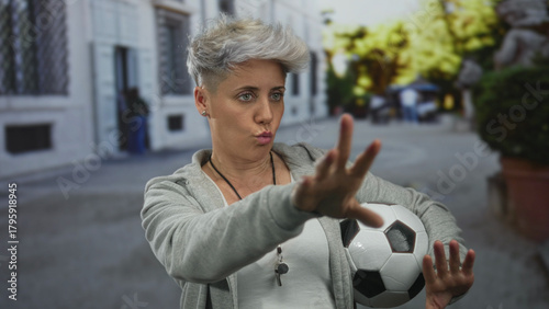 Foto Woman coach in gray hoodie holding soccer ball under arm points finger forward on street; authority discipline motivation leadership