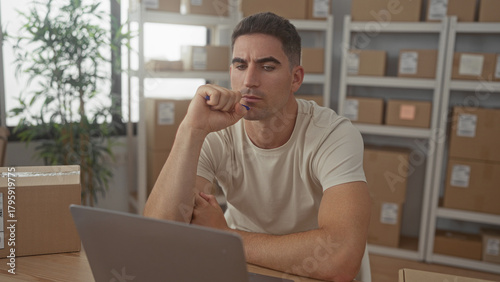 Man with pen to chin and parcel boxes at desk reviewing laptop in office building; contemplation planning clarity.