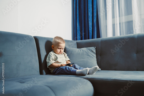 Curious toddler explores smartphone while sitting comfortably on a blue couch in a sunlit living room