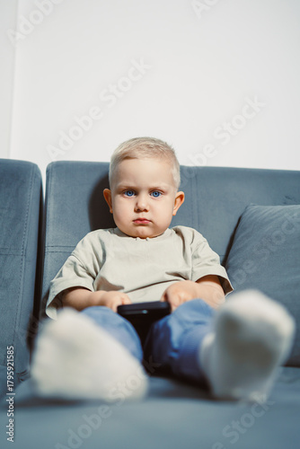 Focused child sitting on a couch while playing with a smartphone in a bright living room during daytime