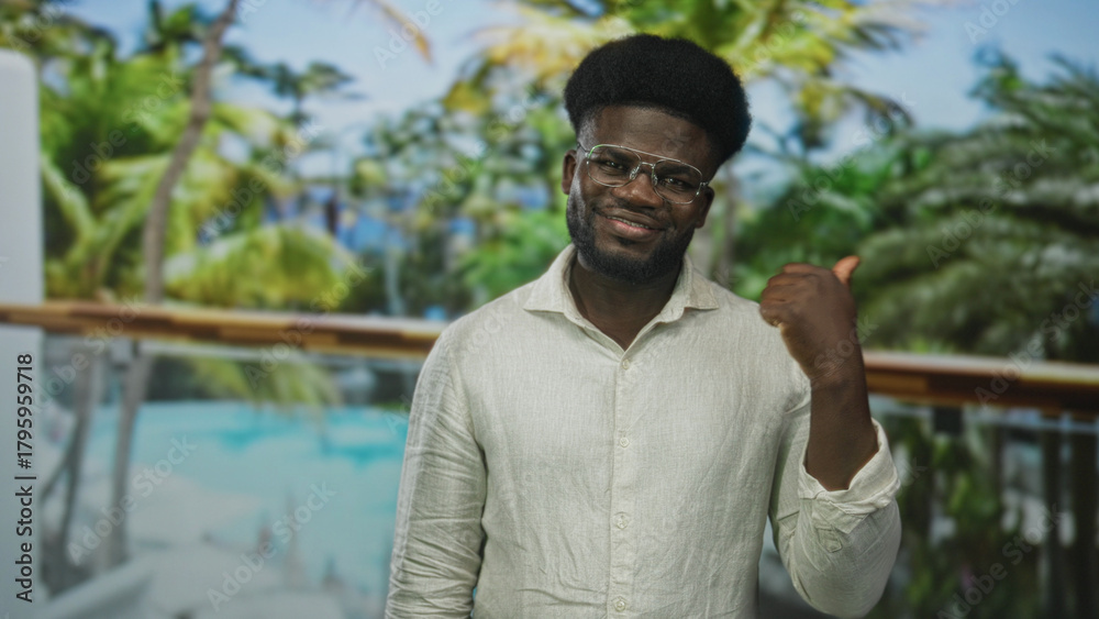 Fototapeta premium Man pointing thumb toward pool and resort building, bare hand raised in a thumbs gesture while smiling and wearing glasses; vacation confidence.