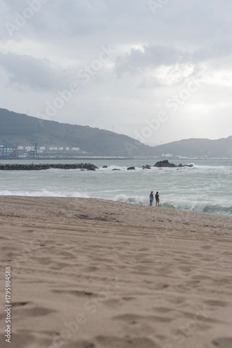 Zwei Personen stehen am Rand eines sandigen Strandes vor einer rauen See, im Hintergrund sind Hügel, Industrieanlagen und ein bewölkter Himmel mit diffusem Licht zu sehen. Die Atmosphäre wirkt ruhig u