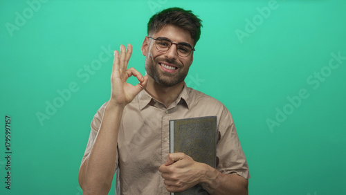 Quadro em tela Smiling man wearing glasses holds book and makes ok gesture with hand in bright green studio; approval