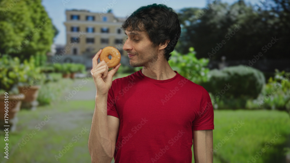 Fototapeta premium Young man smiles while holding a glazed donut near mouth at building entrance on a sunny day; happiness.