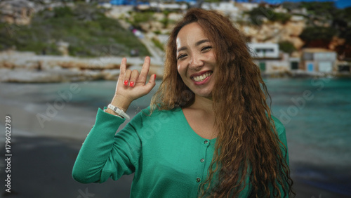 Foto Woman makes rock horns with raised hand at beach, smiling in green sweater with visible hand and red nails; joy summer fun