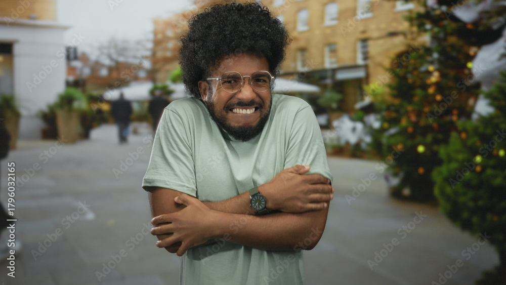 Fototapeta premium Man shivering outdoors on a city street, holding himself in a cold winter breeze, with urban christmas decorations in the background, evoking a chilly, festive atmosphere.