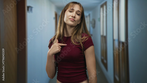 Young woman points finger to chest in hotel building corridor with closed eyes and slight smirk; confidence self belief. Free Download Young woman points finger to chest in hotel building corridor with closed eyes and slight smirk; confidence self belief. - GFXLoad
