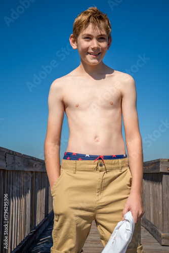 Smiling shirtless boy standing casually outside on a wooden deck during his beach vacation