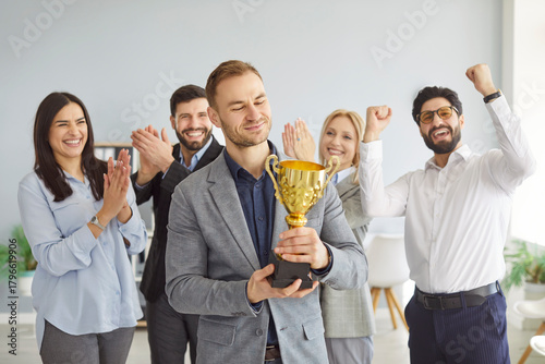 фотографія Happy businessman won prize or trophy, being acknowledged by friendly coworkers, holding golden cup, achieved great success in business sphere, cheerful workers applauding in background