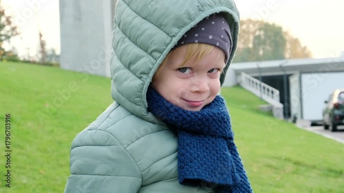A four-year-old boy in a green jacket and blue scarf looks into the camera with a mischievous smile. Perfect 4K video for family, childhood, and lifestyle content.