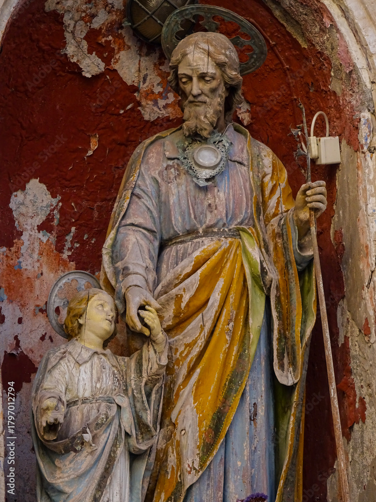 Fototapeta premium Old wooden religious sculpture showing a bearded man leading a haloed child by the hand, with cracked paint and faded colors, photographed in Syracuse, Sicily, Italy.
