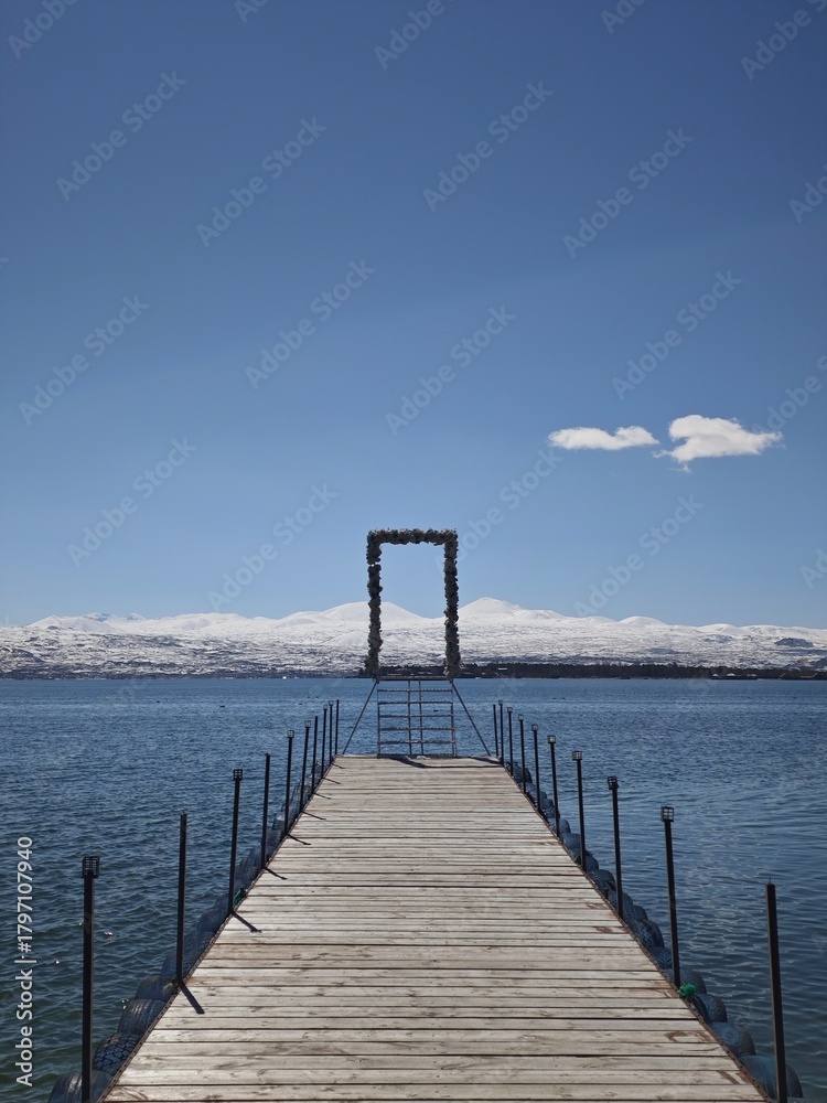 Obraz premium a wooden pier on a calm mountain lake or sea, set against snow-capped peaks and a clear sky.