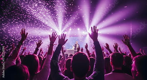 A crowd of people with raised hands enjoying a concert with bright purple stage lights and falling confetti.