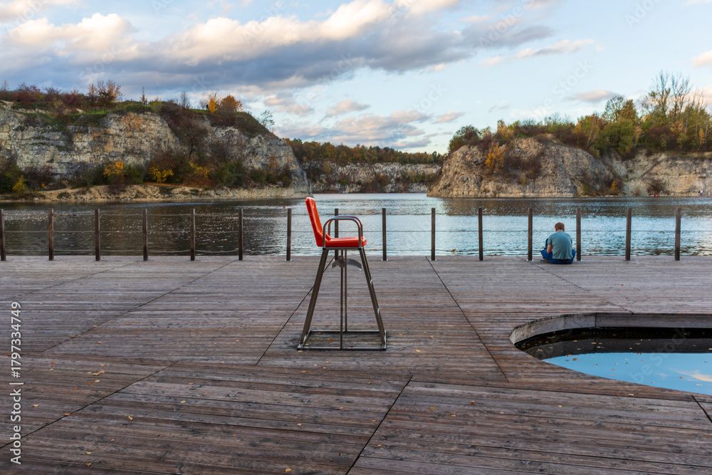 Fototapeta premium Empty red lifeguard chair stands on a wooden lakeside platform overlooking calm water and rocky cliffs. Soft afternoon light and still reflections create a minimal yet atmospheric scene of solitude an