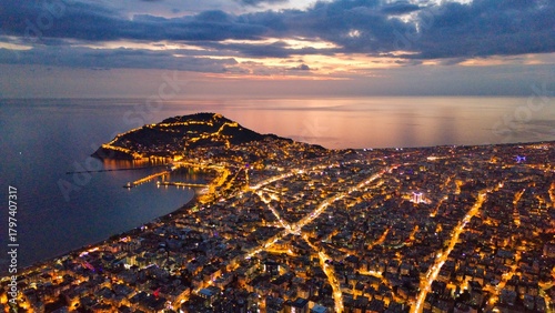 A drone-captured dusk panorama of Alanya, Turkey, showing the illuminated city, peninsula and harbor against a calm sea and twilight sky