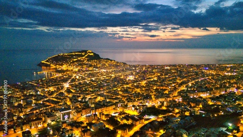 A drone-captured dusk panorama of Alanya, Turkey, showing the illuminated city, peninsula and harbor against a calm sea and twilight sky