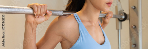 A fit and firm healthy young Asian fitness woman in a gym, performing a barbell behind the neck press exercise for shoulder strength and muscle development, arm closeup view, header banner crop