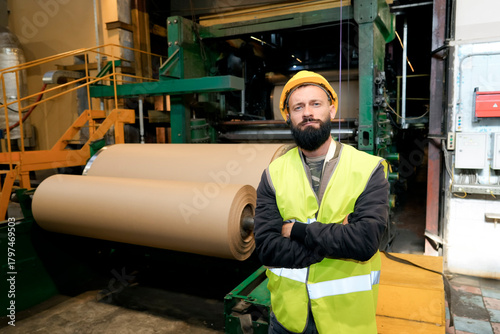In a recycling plant, a man with a beard wearing a helmet and safety vest stands near a machine. He oversees the production of cardboard rolls, ensuring quality in the workshop