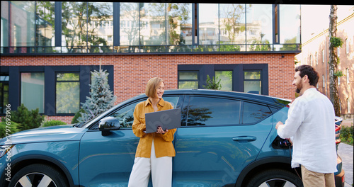 Businessman Charging electric car while businesswoman works on laptop outdoors. Eco-friendly technology and modern lifestyle