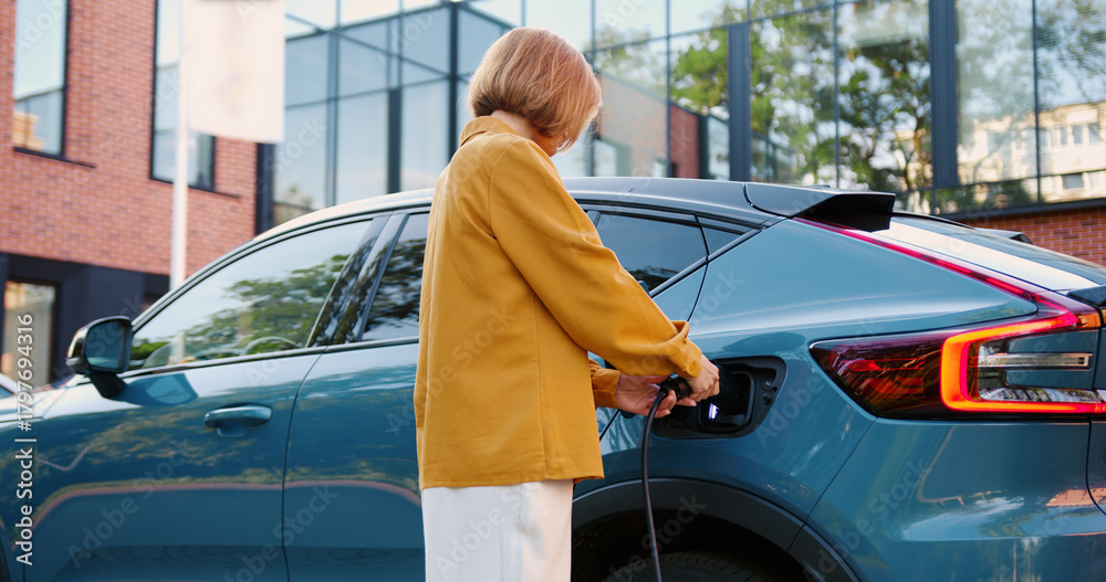 Fototapeta premium Modern stylish woman connects charging cable parked electric vehicle. Concept of eco transport, green energy, and modern lifestyle.