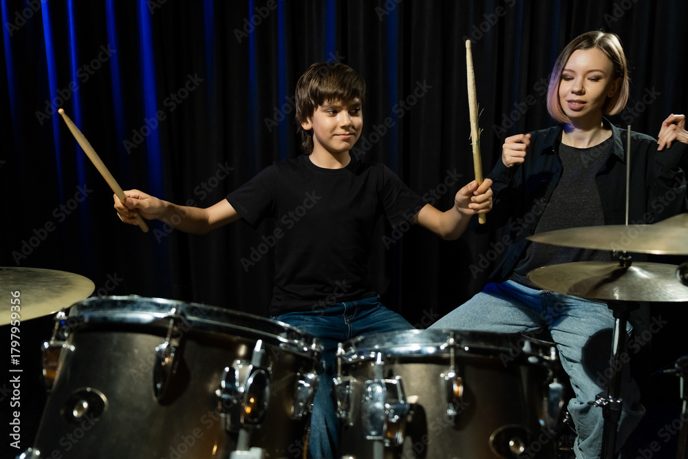 Fototapeta premium Young caucasian woman teaches a boy to play the drums in the studio on a black background. Music school student