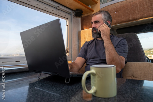 A bearded man uses his laptop inside a camper van while talking on a mobile phone, with bright daylight coming through the window and a mug on the table.