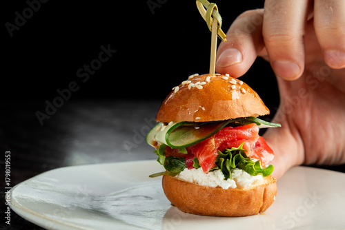 A hand holds a gourmet mini burger with salmon, cream cheese, tomato, and cucumber on a sesame seed bun, secured by a skewer on a white plate