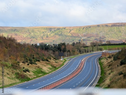 An elevated wide shot captures a modern, dual-carriageway highway curving through a rolling, semi-rural landscape under a pale, overcast sky. The road is flanked by grassy slopes and sparse trees