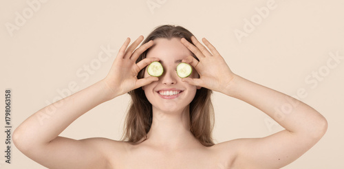 Beauty, health care and cosmetology concept. Beautiful young woman covering her eyes with cucumber slices. Happy model with smiling face against beige background with copy space