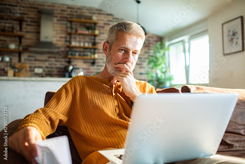 Canvas Print Mature man reviewing bills on laptop at home, thoughtful