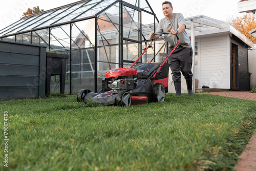 Man mowing narrow backyard lawn beside modern metal fence and garden plants.