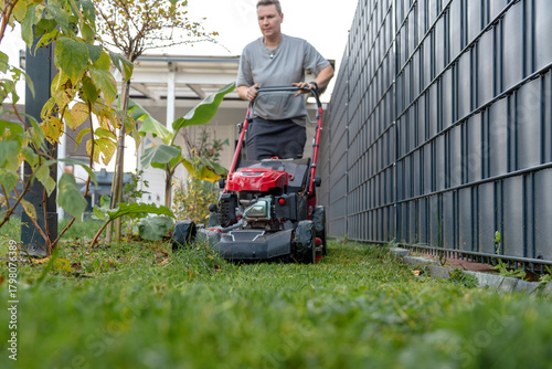 Man mowing narrow backyard lawn beside modern metal fence and garden plants.