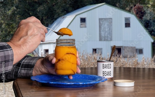 A person is spooning out peaches from a jar while sitting at a table. In the background, an old barn is visible among the trees. A can of baked beans sits next to the jar.
