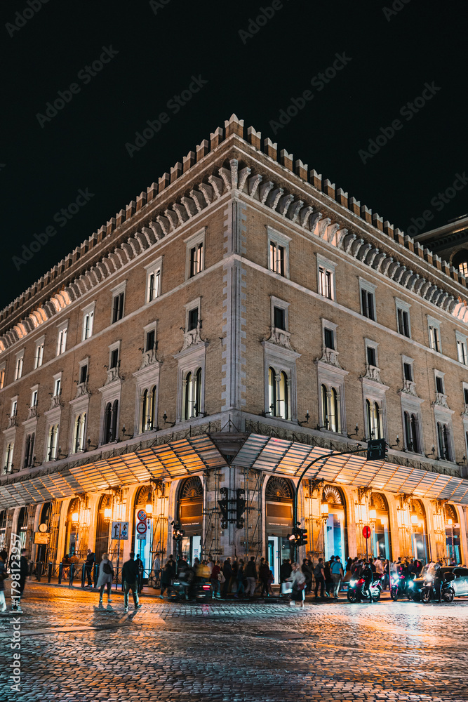 Naklejka premium Exterior corner view of a large, well-lit historic building on a cobblestone street at night with people gathered near the ground floor.